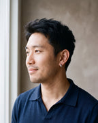 Man in a navy fine-gauge knit polo with an open collar by a window wearing the silver-tone Atlas 4mm hoop earrings