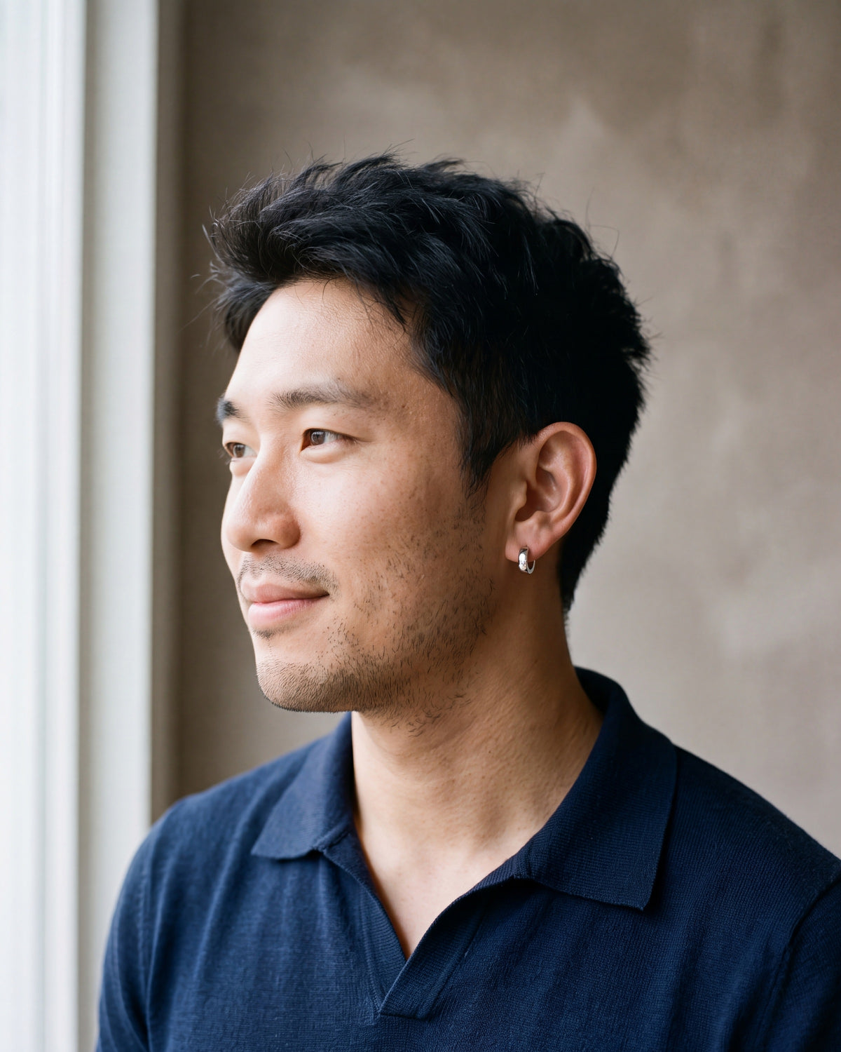 Man in a navy fine-gauge knit polo with an open collar by a window wearing the silver-tone Atlas 4mm hoop earrings