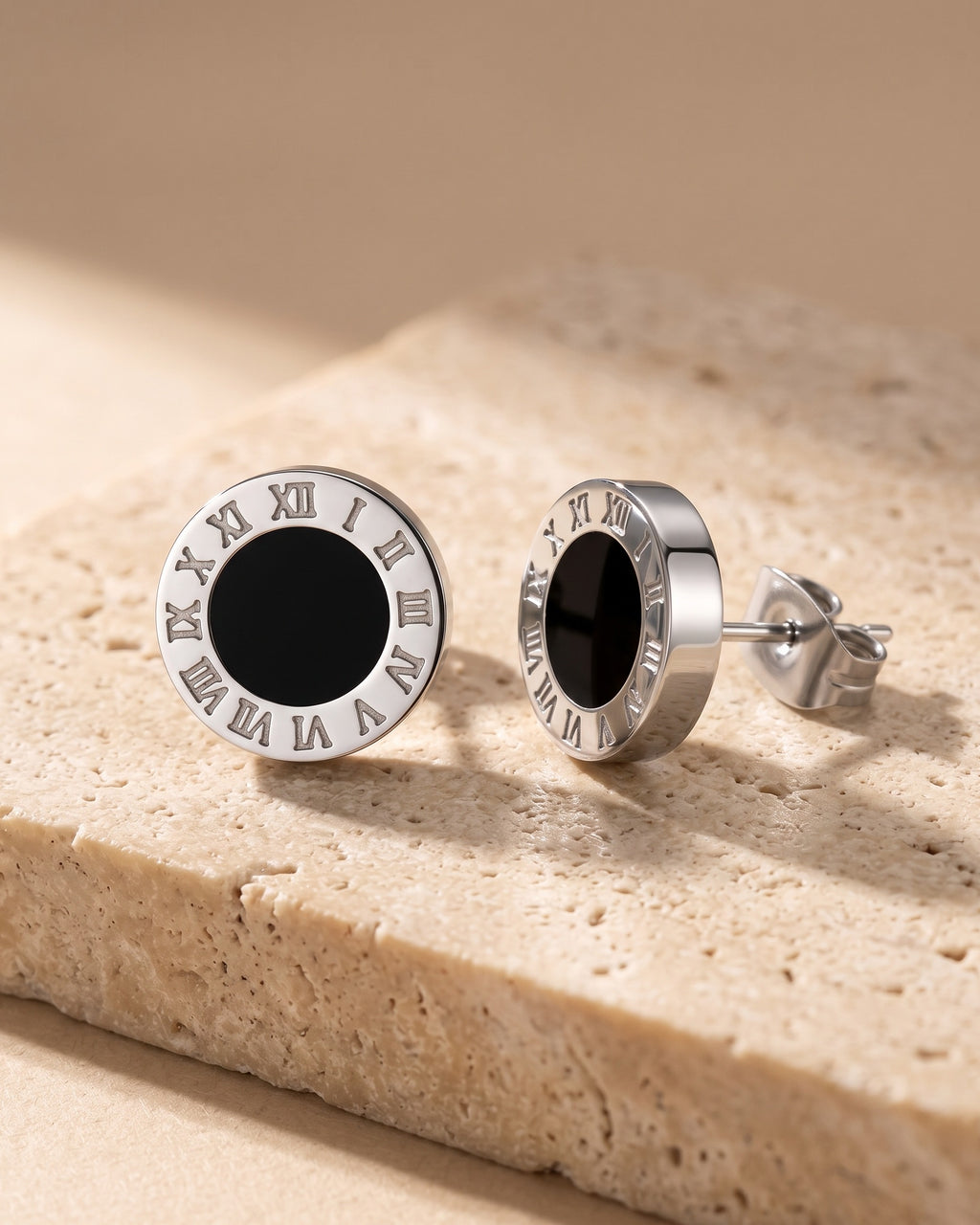The pair of silver-tone Atlas 8mm Roman numeral stud earrings with black center on warm travertine stone