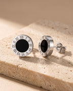 The pair of silver-tone Atlas 8mm Roman numeral stud earrings with black center on warm travertine stone