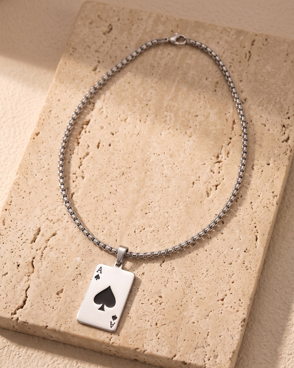 The whole silver-tone Atlas Ace of Spades Pendant Necklace lying on warm travertine stone
