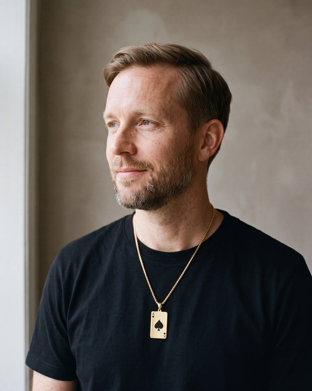 Man in a heavyweight black cotton crew-neck t-shirt by a window wearing the gold-tone Atlas Ace of Spades Pendant Necklace