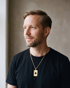 Man in a heavyweight black cotton crew-neck t-shirt by a window wearing the gold-tone Atlas Ace of Spades Pendant Necklace