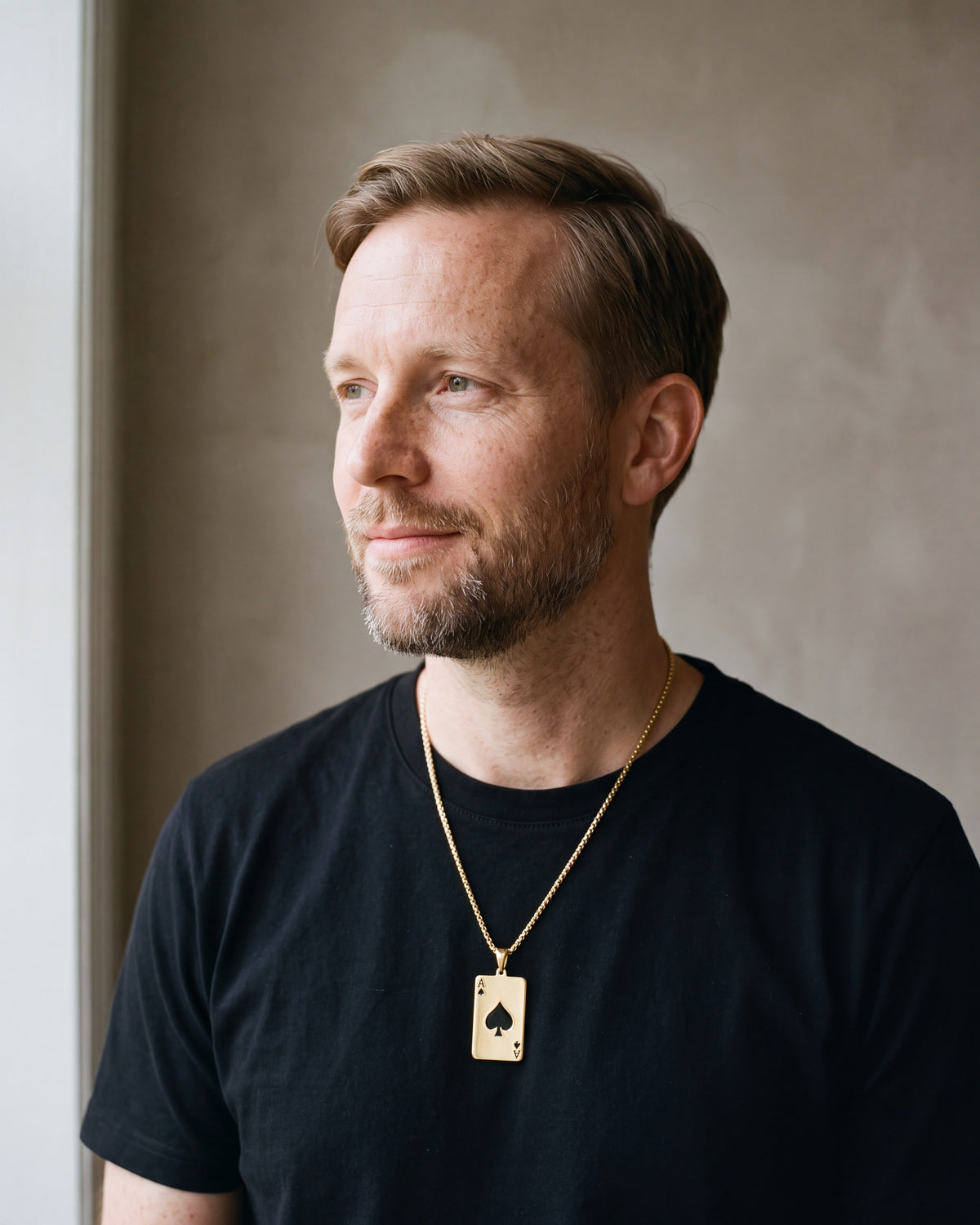 Man in a heavyweight black cotton crew-neck t-shirt by a window wearing the gold-tone Atlas Ace of Spades Pendant Necklace