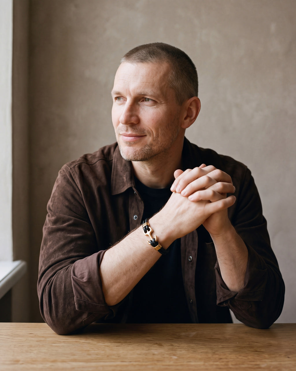 Man in a brown suede overshirt seated at a wooden table, wearing the gold-tone Atlas anchor bracelet