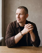Man in a brown suede overshirt seated at a wooden table, wearing the gold-tone Atlas anchor bracelet