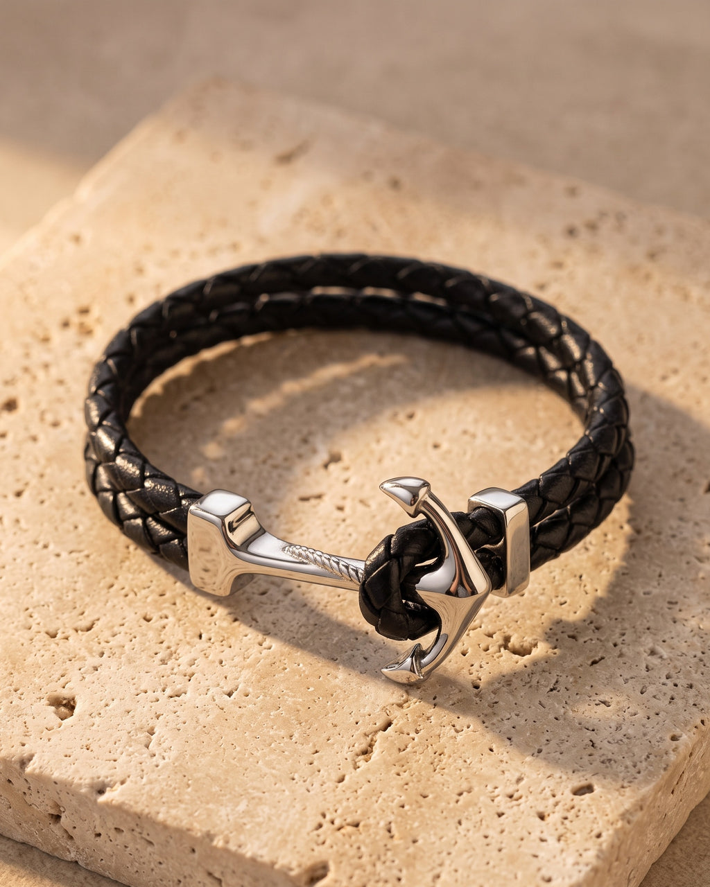 Entire Atlas anchor hook bracelet in braided leather in black steel, lying flat on warm stone