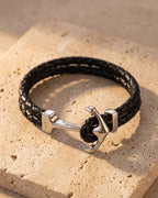 Entire Atlas anchor hook bracelet in braided leather in black steel, lying flat on warm stone