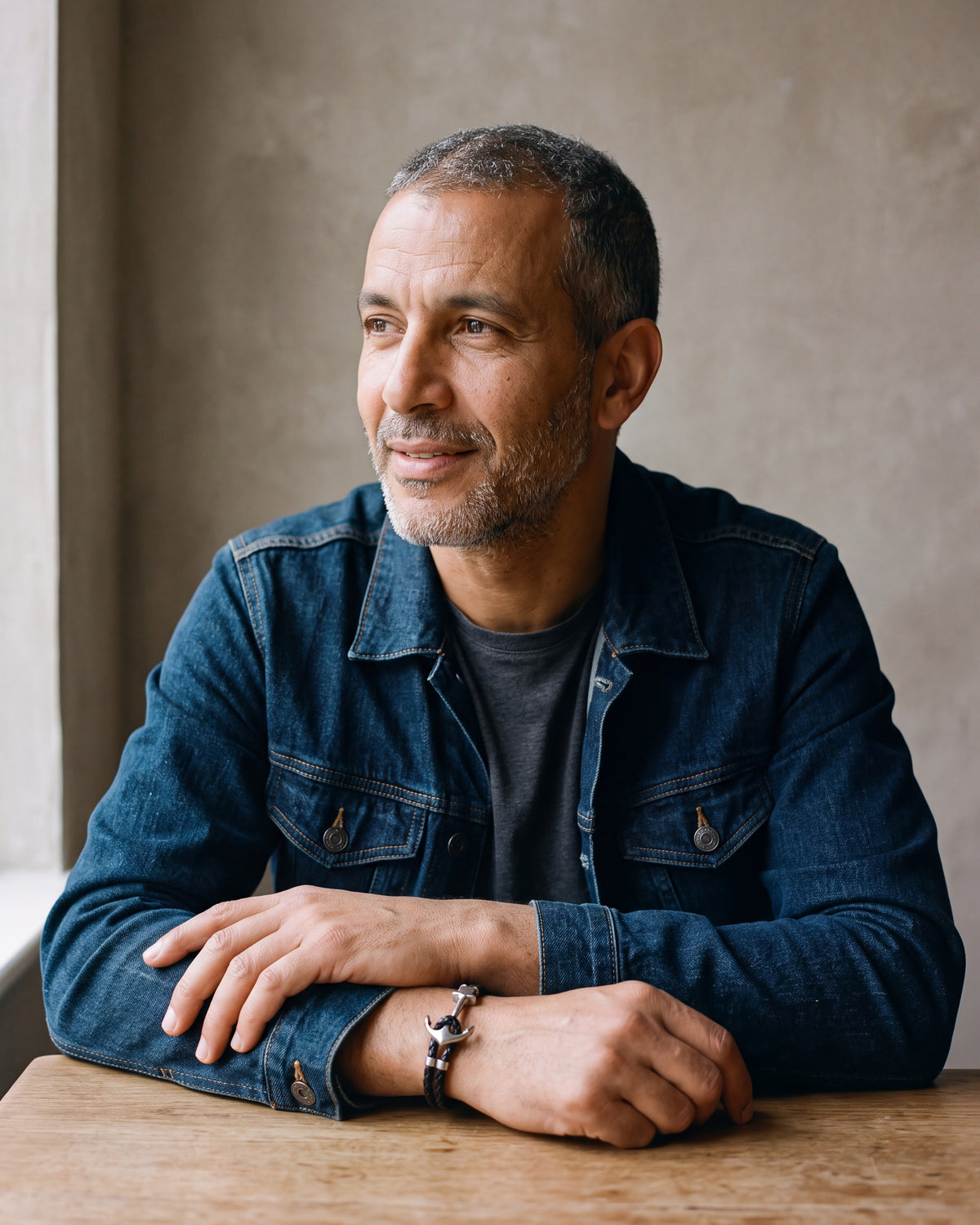 Man in a denim jacket over a charcoal t-shirt by a window wearing the Atlas anchor hook bracelet in braided leather in black steel