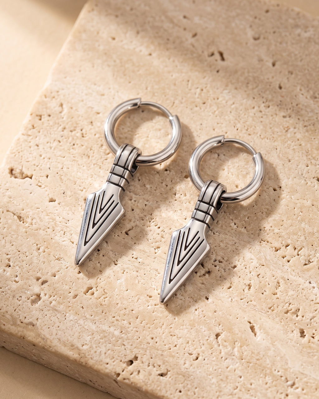 Entire Atlas arrowhead dangle hoop earrings in silver-tone steel, lying flat on warm stone