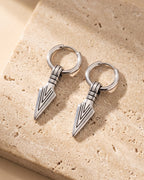 Entire Atlas arrowhead dangle hoop earrings in silver-tone steel, lying flat on warm stone