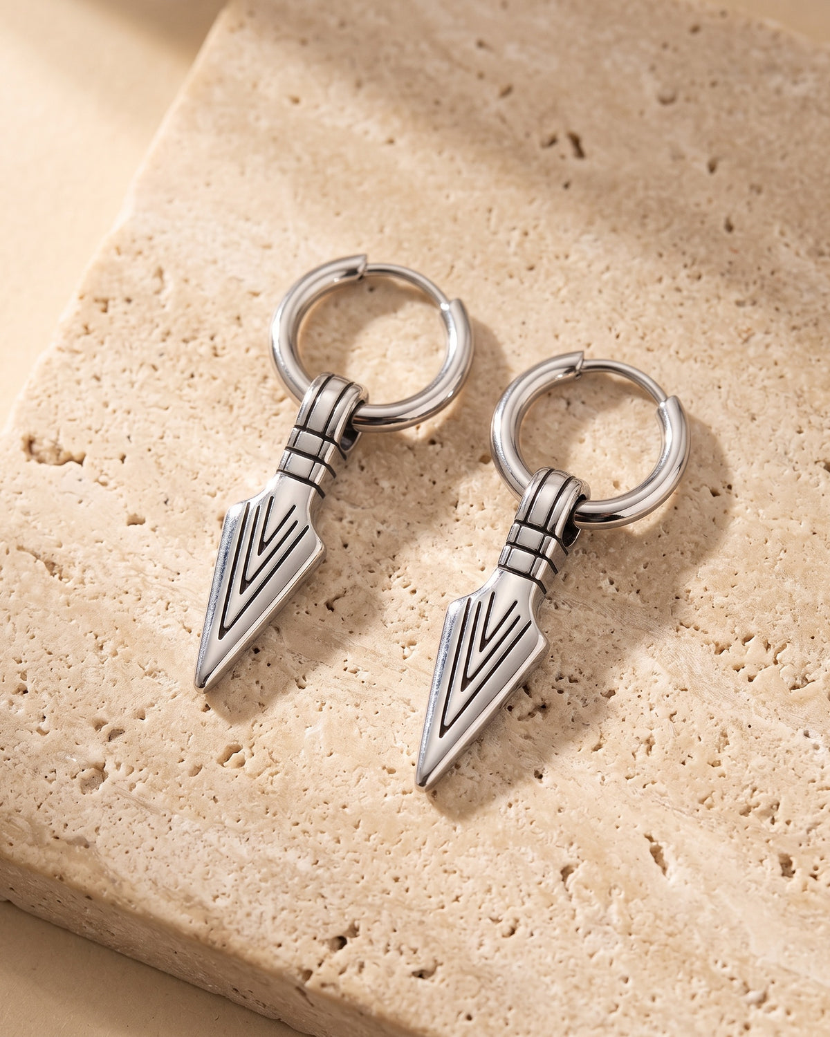 Entire Atlas arrowhead dangle hoop earrings in silver-tone steel, lying flat on warm stone