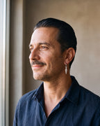 Man in a navy linen shirt by a window wearing the Atlas arrowhead dangle hoop earrings in silver-tone steel