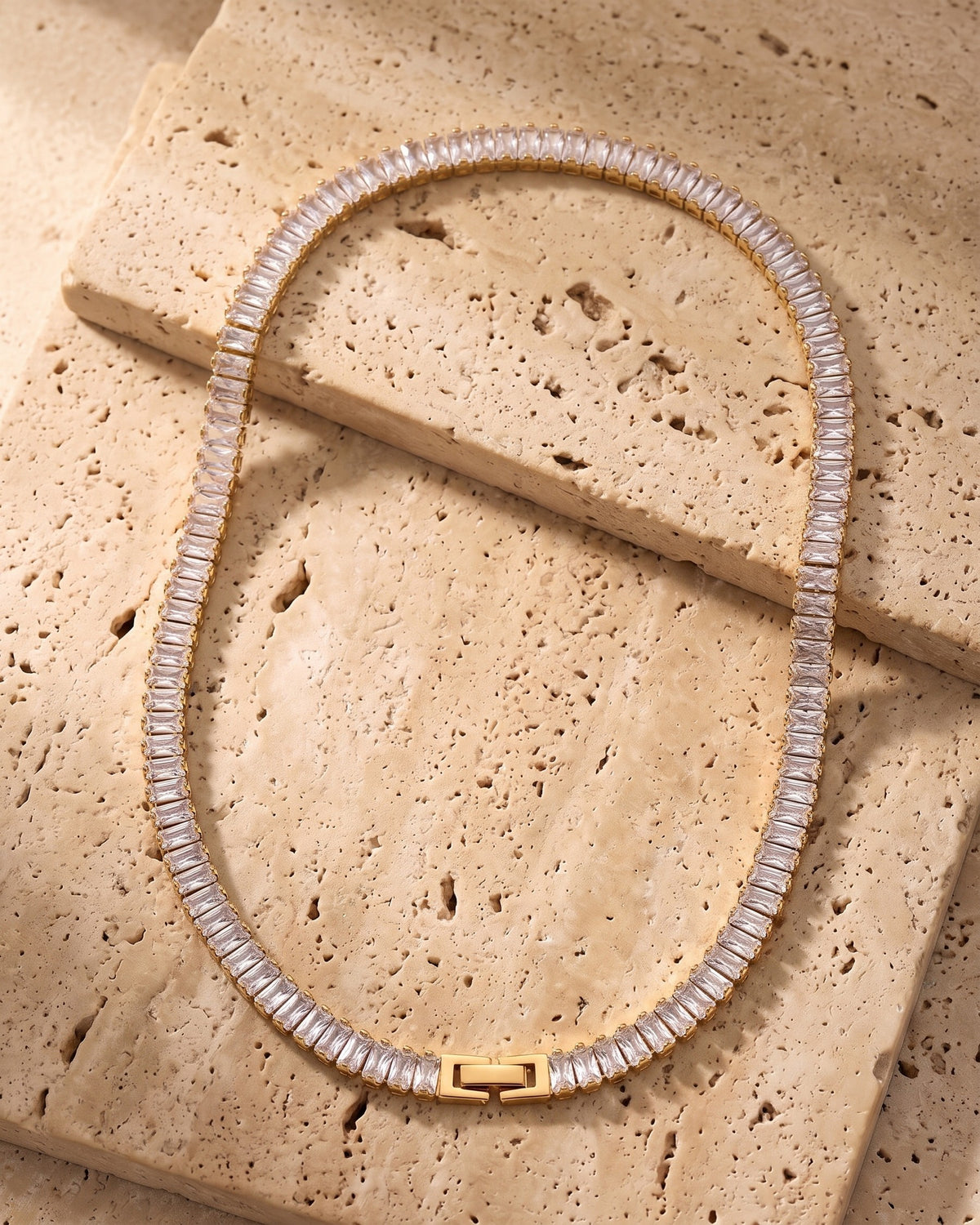 The gold-tone Atlas Baguette Tennis Chain laid on warm travertine stone