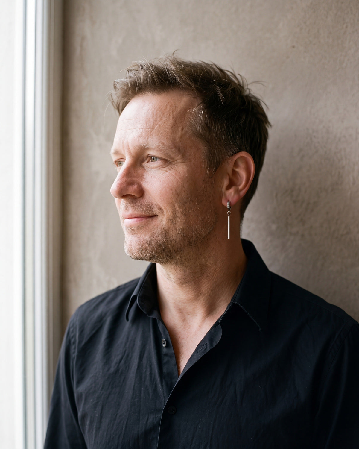 Man in a black fine cotton shirt by a window wearing the silver-tone Atlas bar drop hoop earrings