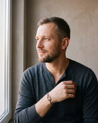 Man in a fine-gauge charcoal-grey merino knit with a henley neckline by a window wearing the silver-tone Atlas box link shackle bracelet