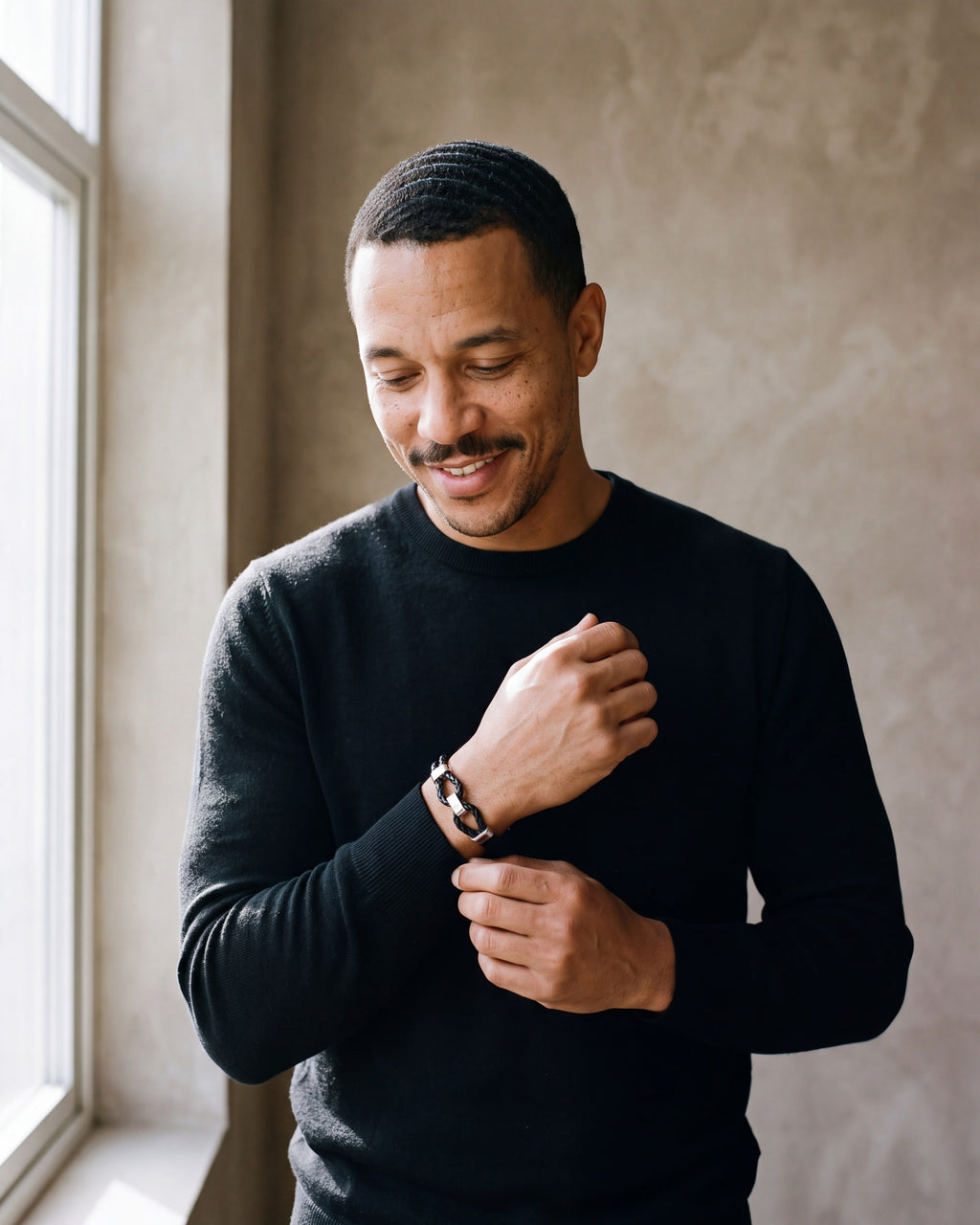 Man in a black merino sweater adjusting his sleeve wearing the black and silver-tone Atlas braided leather shackle bracelet