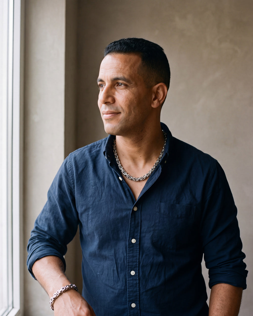 Man in a dark navy cotton oxford shirt worn open at the collar (two buttons open) by a window wearing the silver Atlas byzantine chain and bracelet set