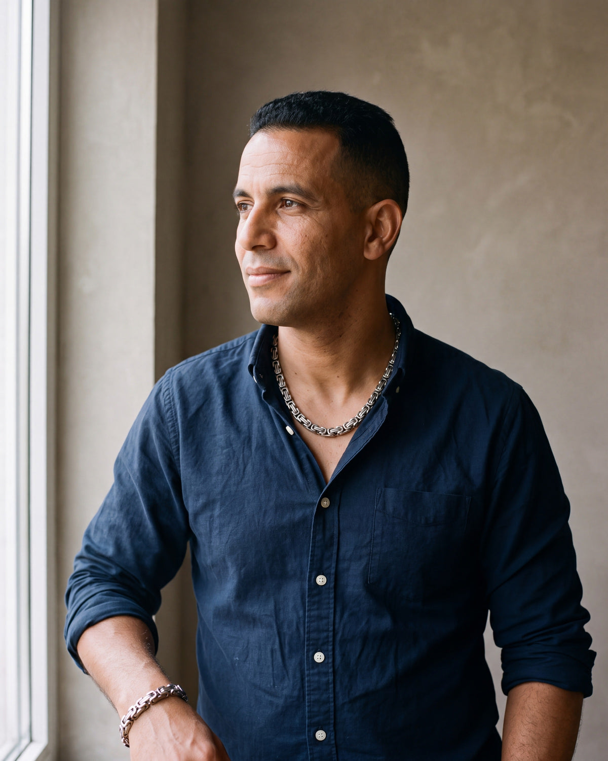 Man in a dark navy cotton oxford shirt worn open at the collar (two buttons open) by a window wearing the silver Atlas byzantine chain and bracelet set