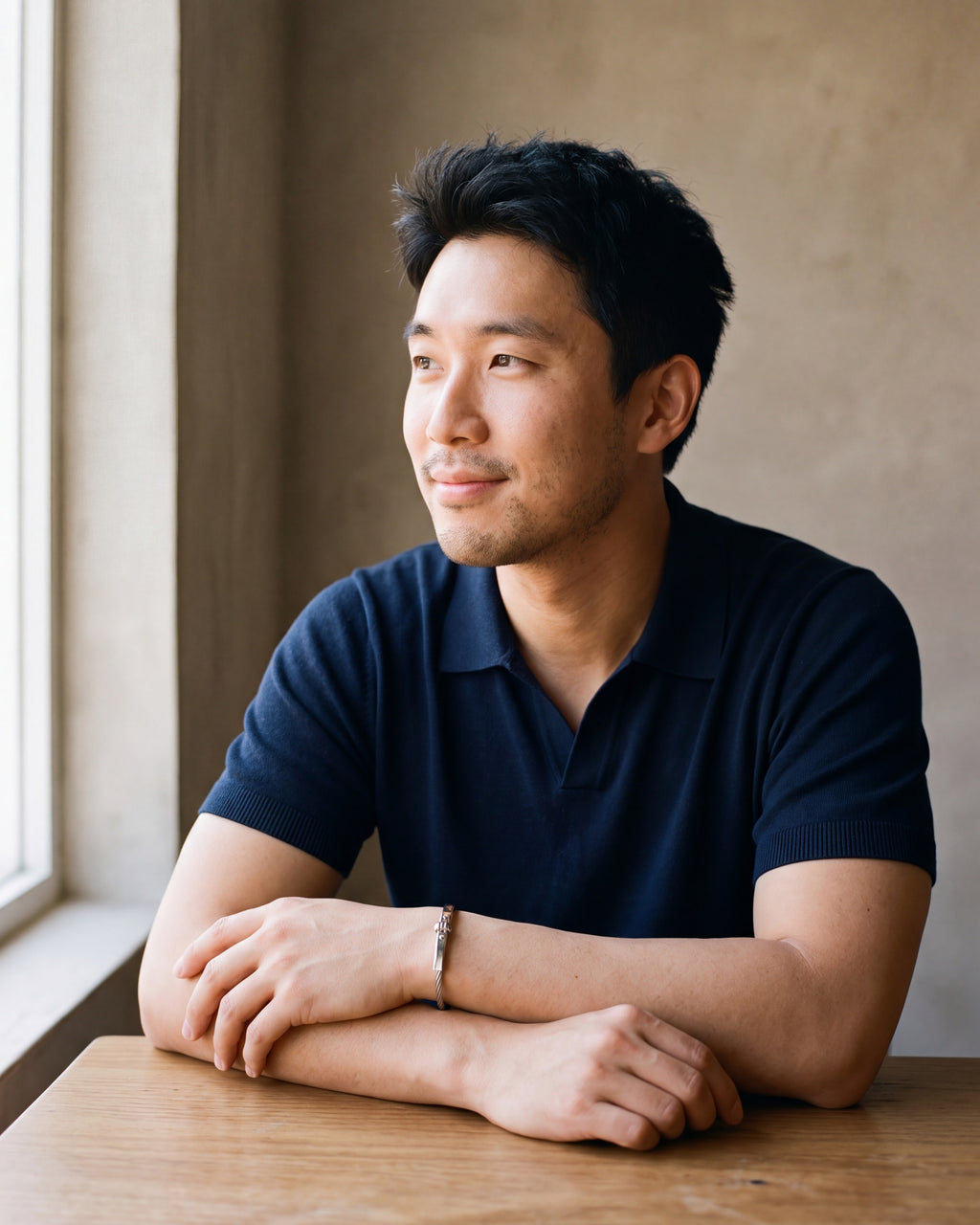 Man in a navy fine-gauge knit polo with an open collar by a window wearing the silver Atlas cable bar bracelet