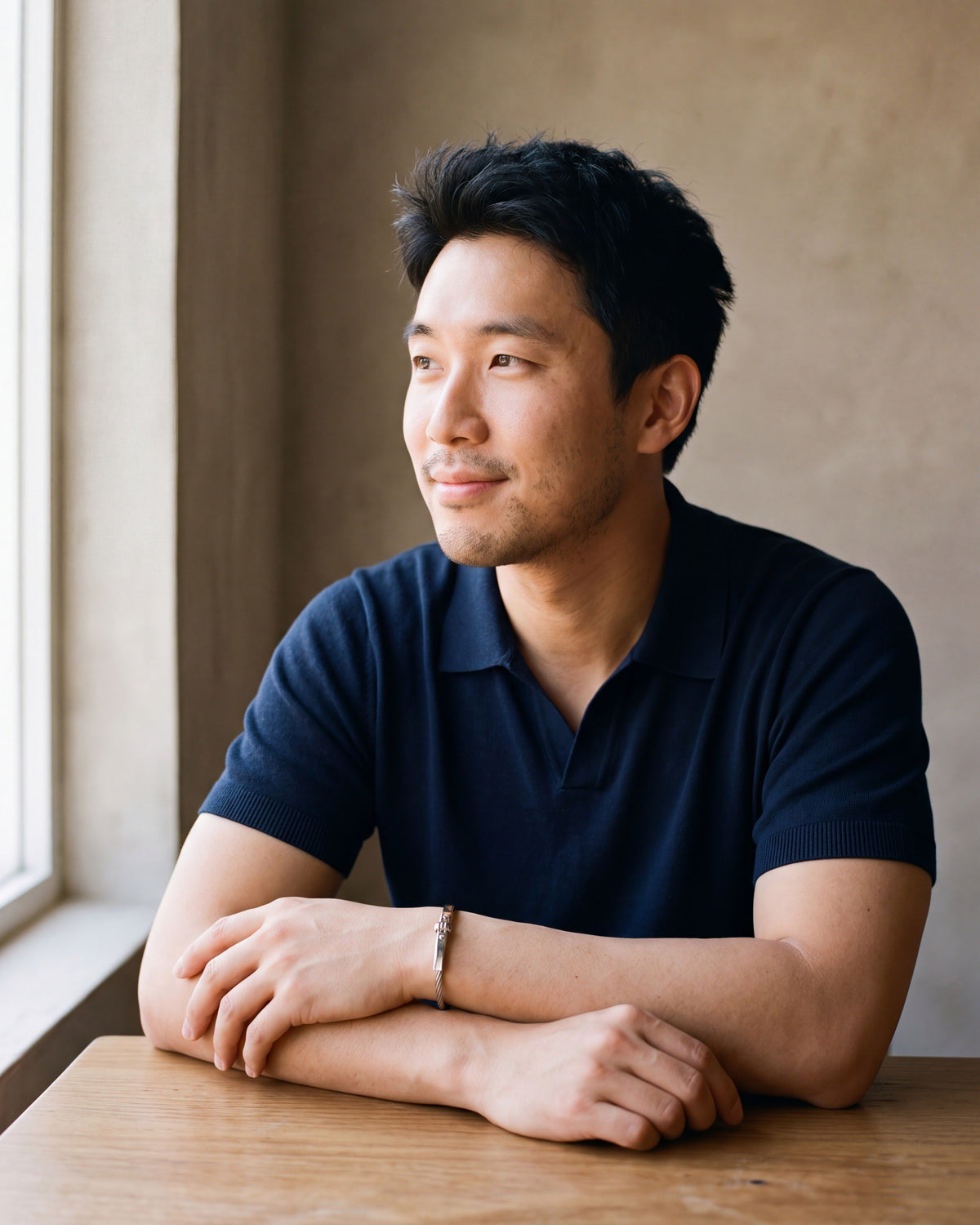 Man in a navy fine-gauge knit polo with an open collar by a window wearing the silver Atlas cable bar bracelet