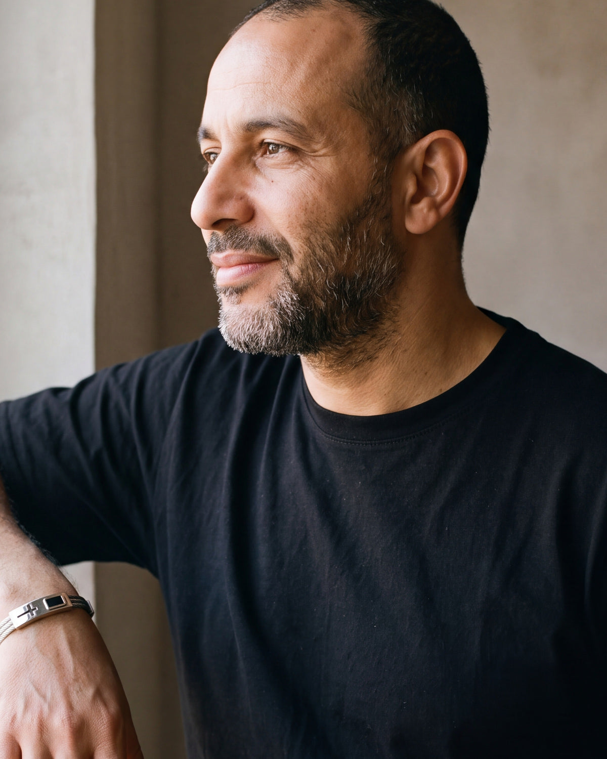 Man in a black t-shirt looking toward the window, wearing the silver Atlas cable cross bracelet