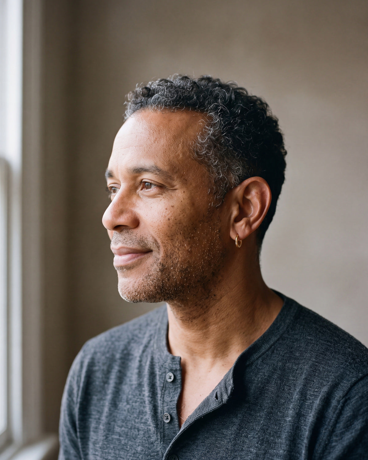 Man in a charcoal henley by a window wearing a gold-tone Atlas classic hoop earring