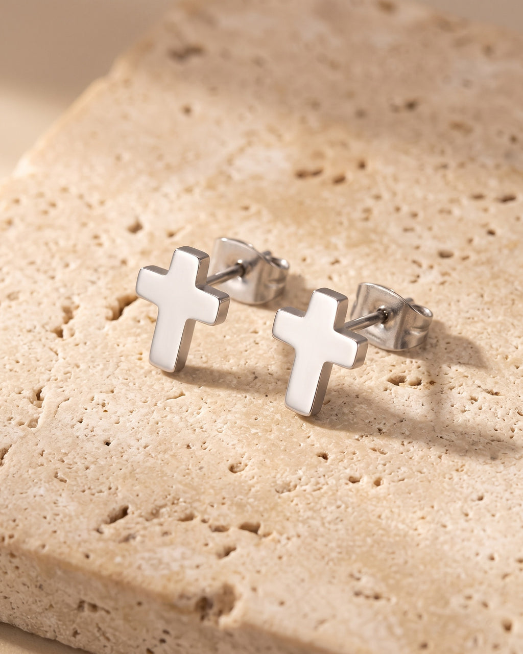 Entire Atlas cross stud earrings in silver-tone steel, lying flat on warm stone