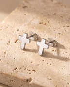 Entire Atlas cross stud earrings in silver-tone steel, lying flat on warm stone