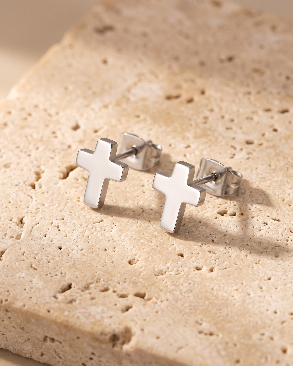 Entire Atlas cross stud earrings in silver-tone steel, lying flat on warm stone
