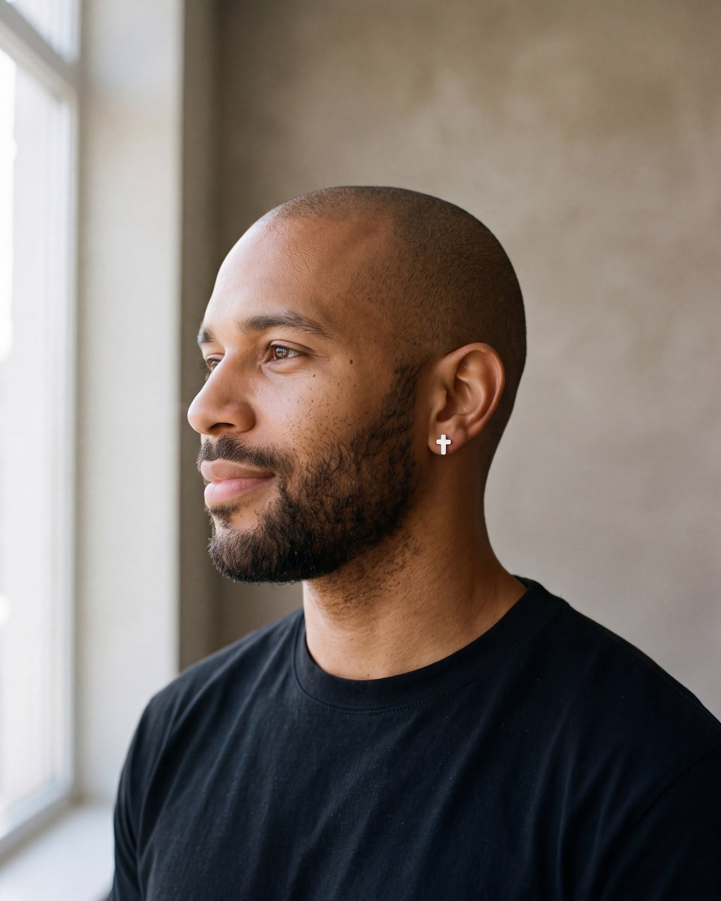 Man in a black t-shirt by a window wearing the Atlas cross stud earrings in silver-tone steel