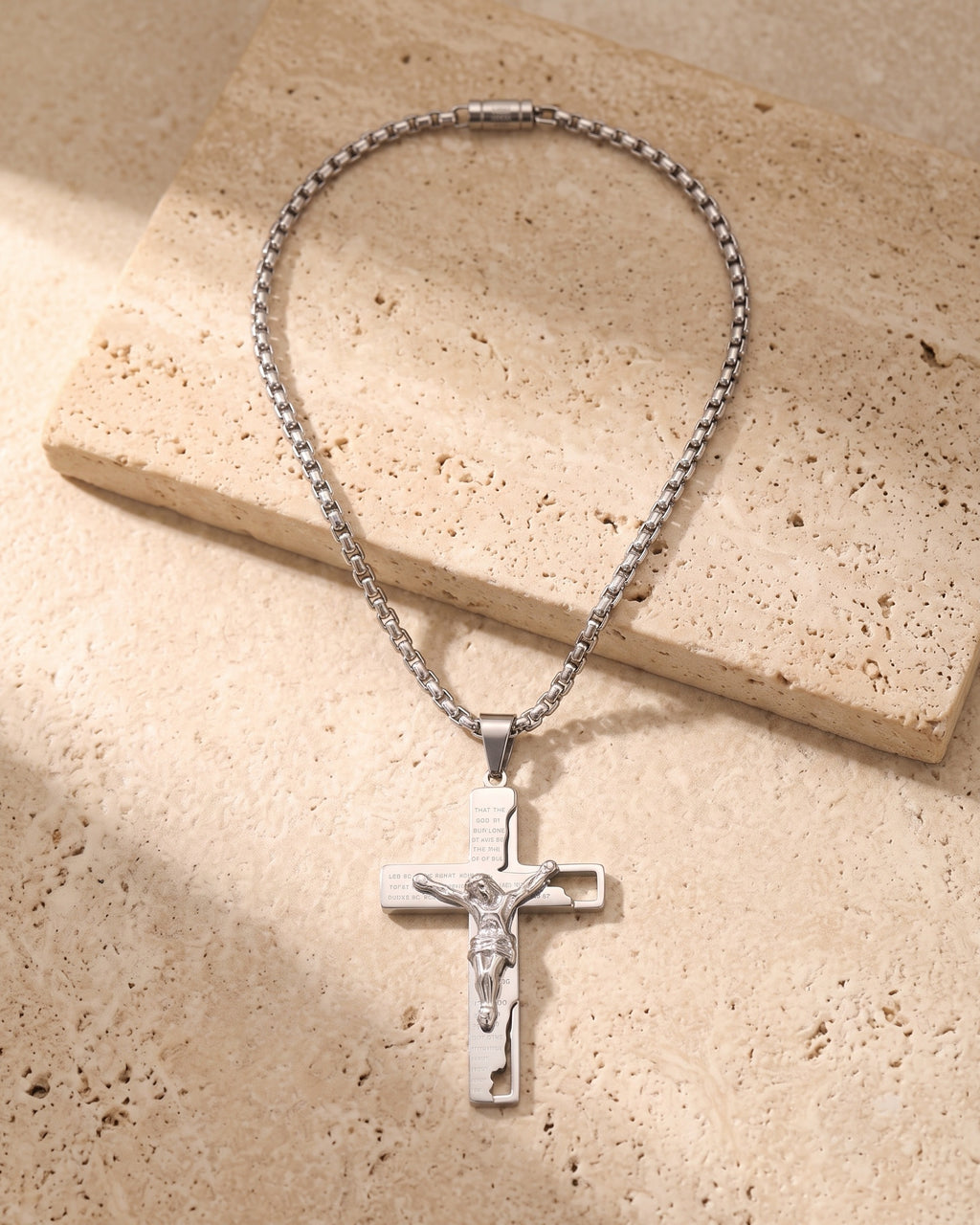 The whole silver-tone Atlas engraved crucifix pendant necklace lying on warm travertine stone