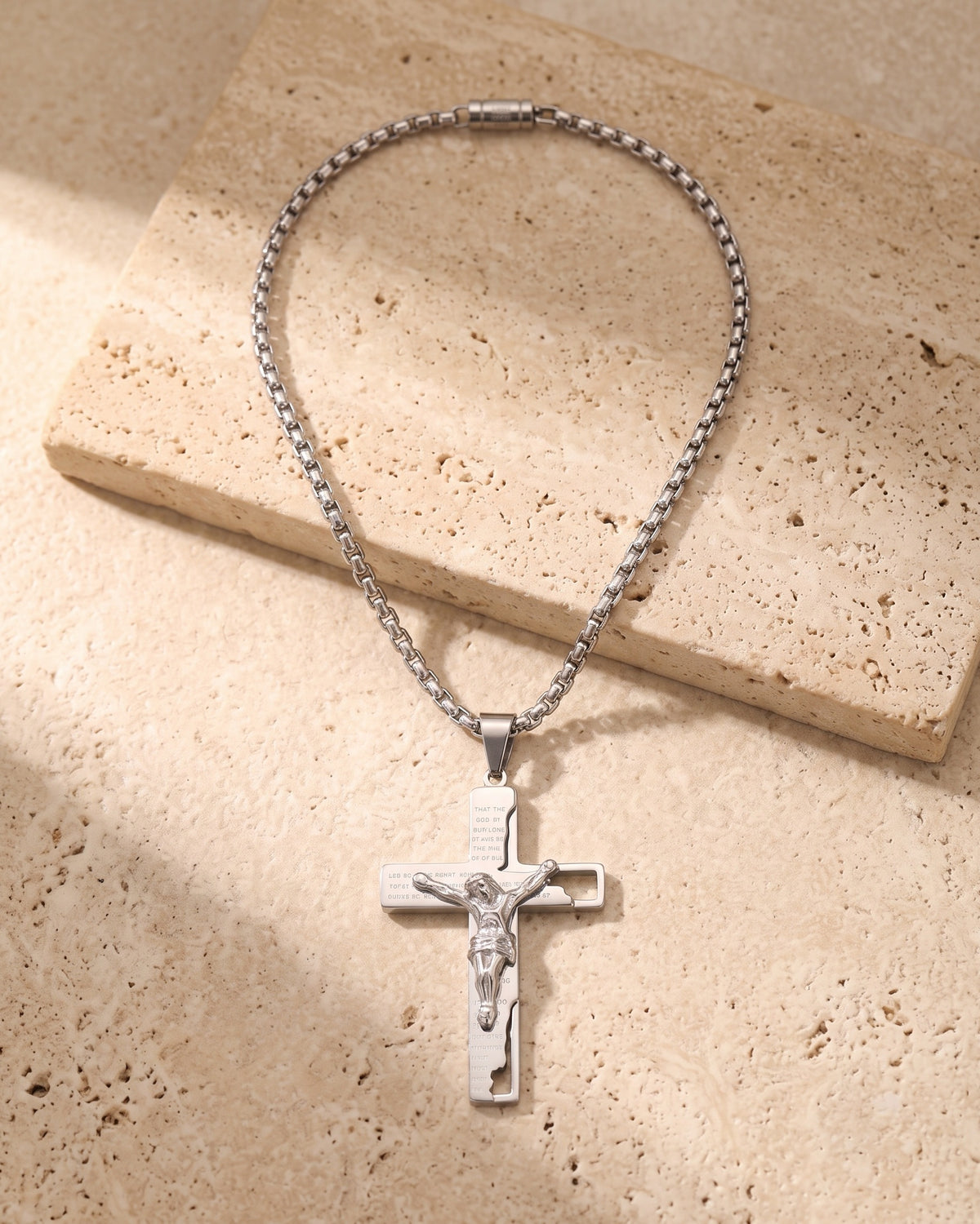The whole silver-tone Atlas engraved crucifix pendant necklace lying on warm travertine stone