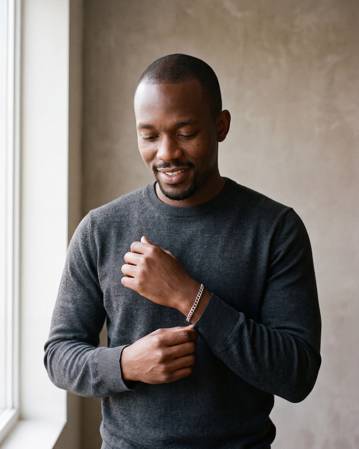 Man in a charcoal knit sweater by a window wearing the Atlas Cuban link bracelet in silver-tone steel