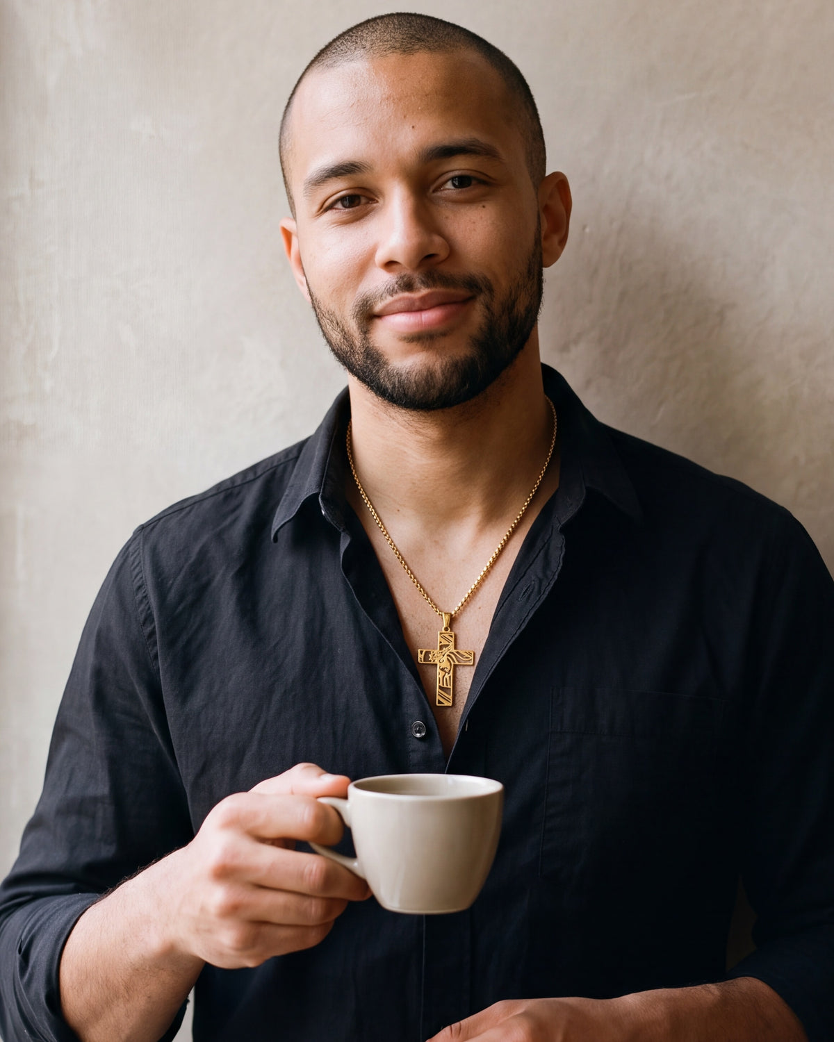 Man in a black shirt holding a coffee cup, wearing the gold-tone Atlas cut-out cross pendant