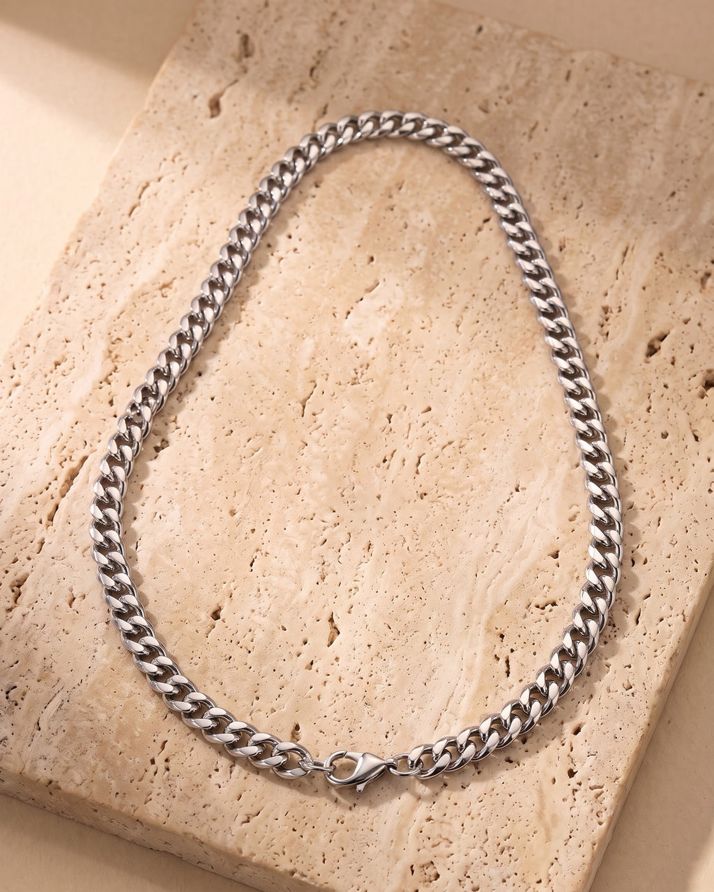 The silver Atlas Dense Cuban Link Chain in Silver-Tone Steel laid on warm travertine stone
