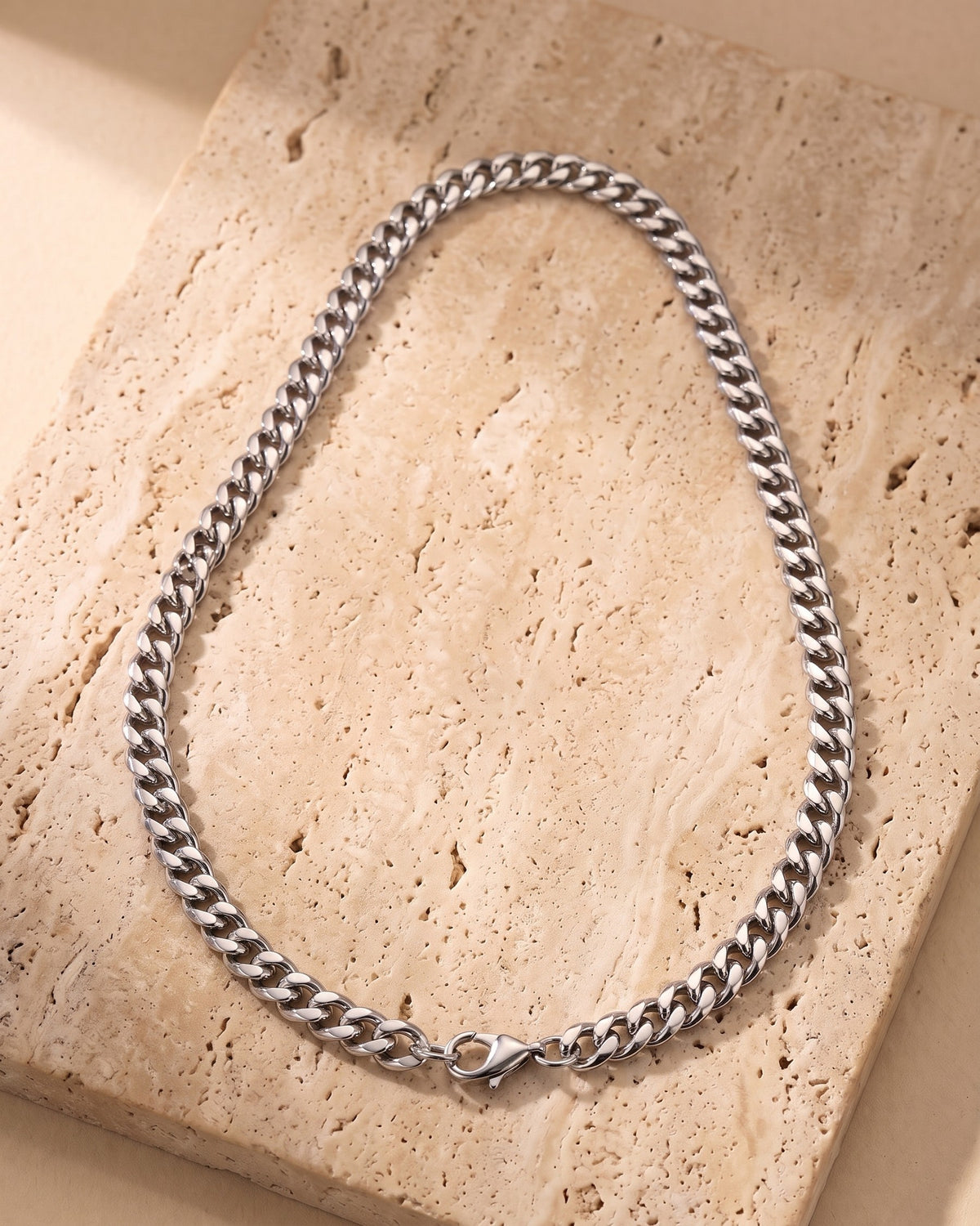 The silver Atlas Dense Cuban Link Chain in Silver-Tone Steel laid on warm travertine stone