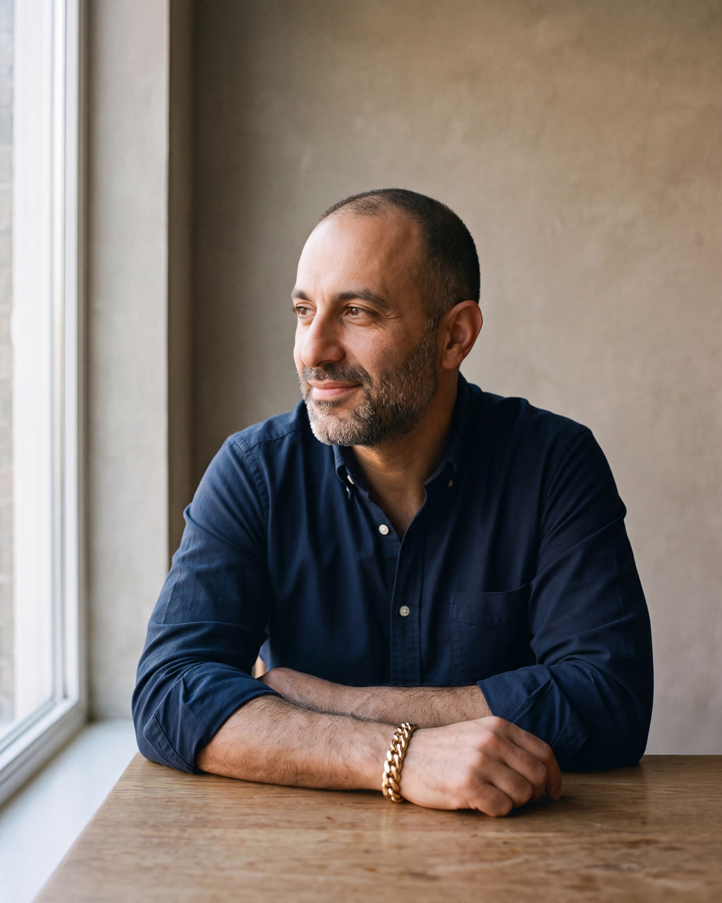 Man in a navy oxford shirt by a window wearing the Atlas double-latch Cuban bracelet in gold-tone steel