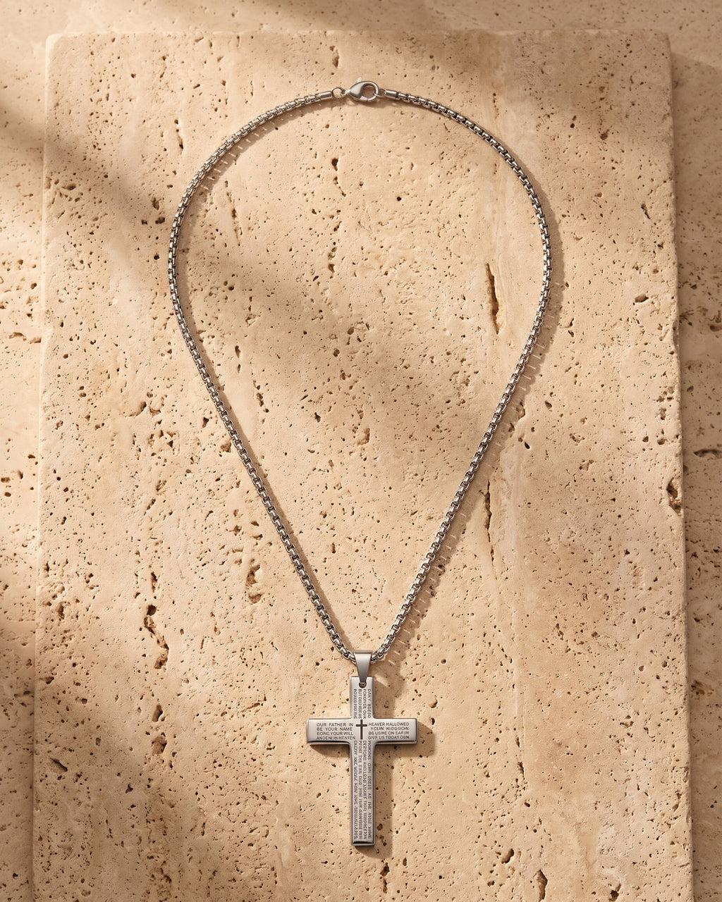 The silver Atlas Engraved Cross Necklace in Titanium Steel laid on warm travertine stone