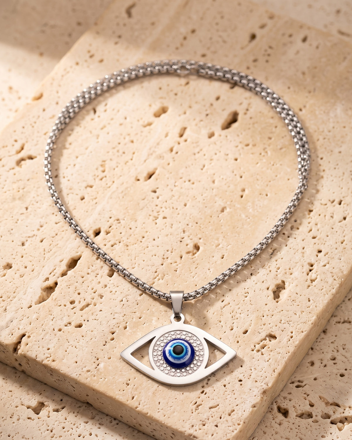 The whole silver Atlas evil eye pendant necklace lying on warm travertine stone