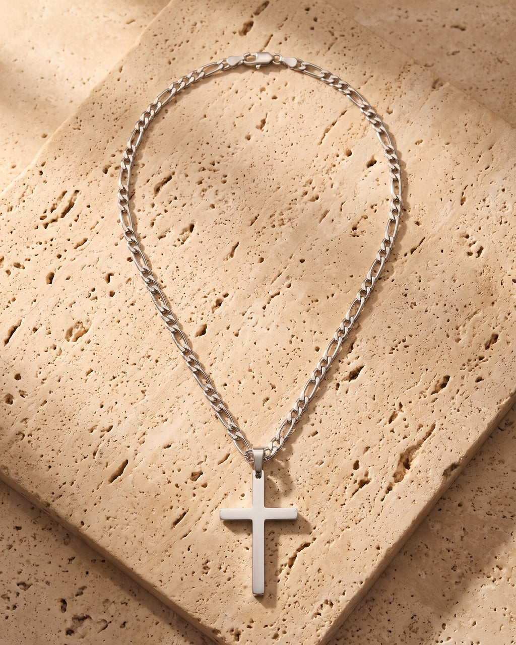 The silver Atlas Figaro Chain Cross Necklace in Titanium Steel laid on warm travertine stone