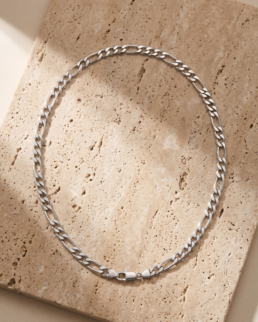 The silver Atlas Figaro Link Chain in Stainless Steel laid on warm travertine stone