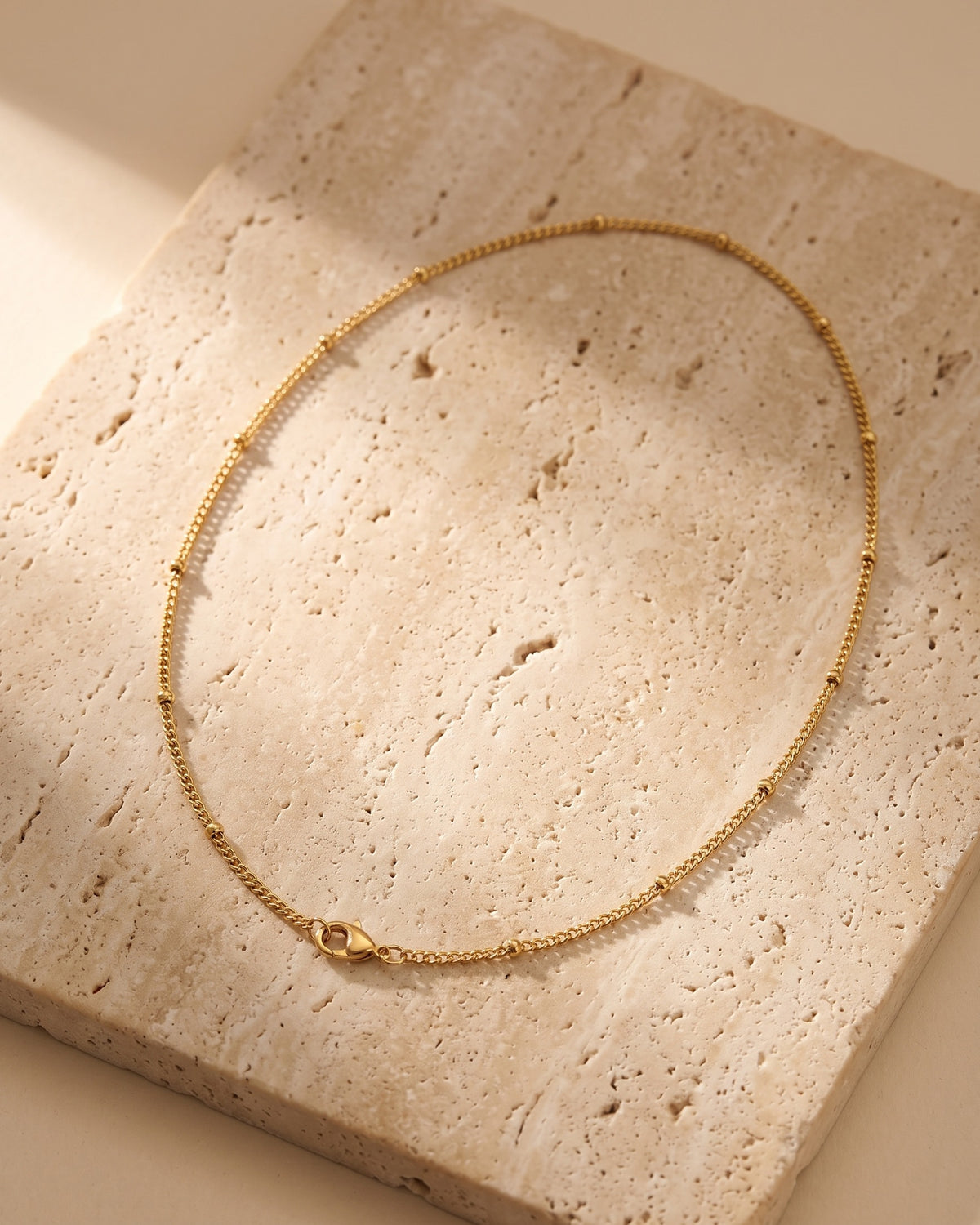 The whole Atlas fine 1 mm bead chain necklace in gold-tone stainless steel laid on warm stone