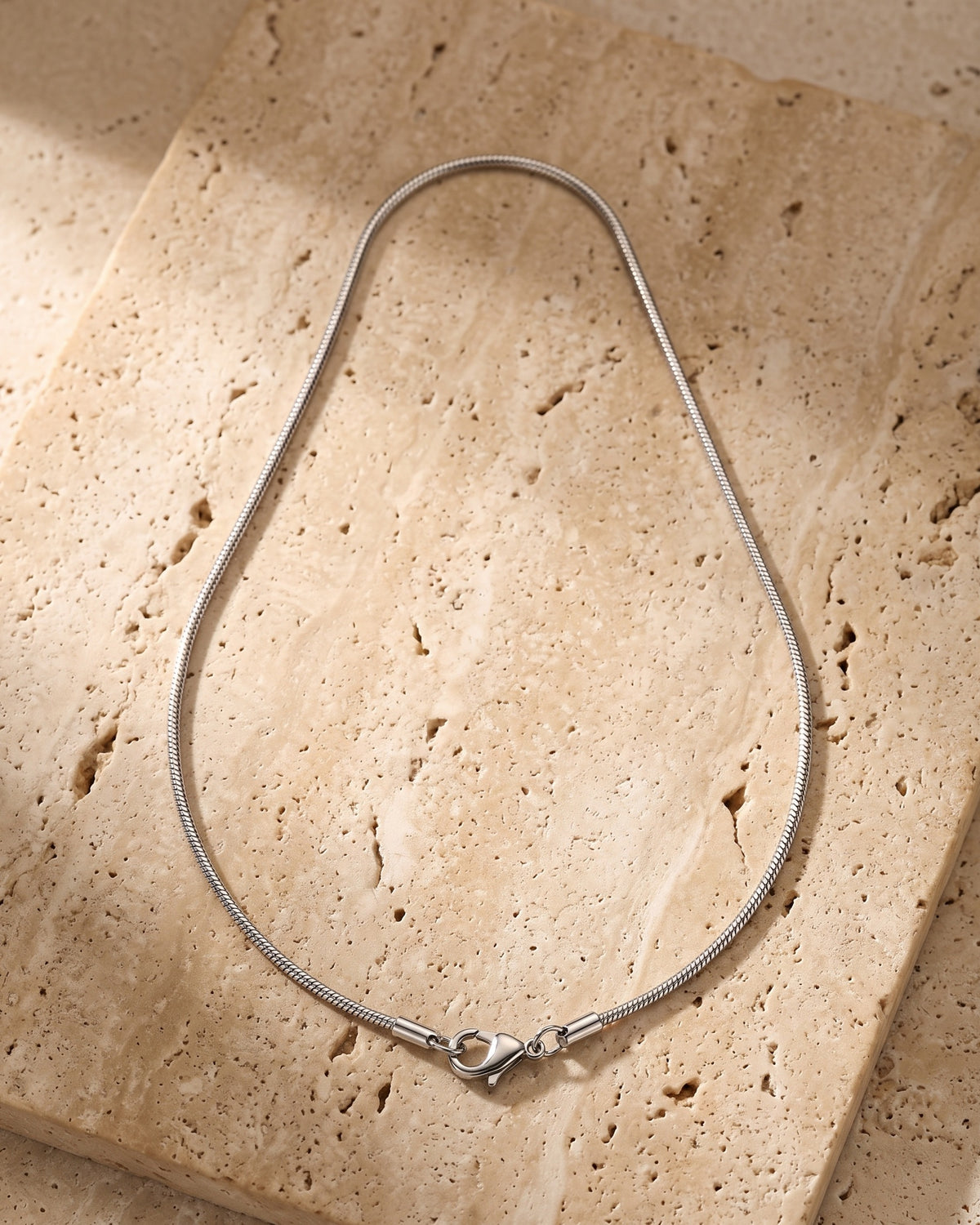 The whole silver-tone Atlas Fine Snake Chain lying on warm travertine stone