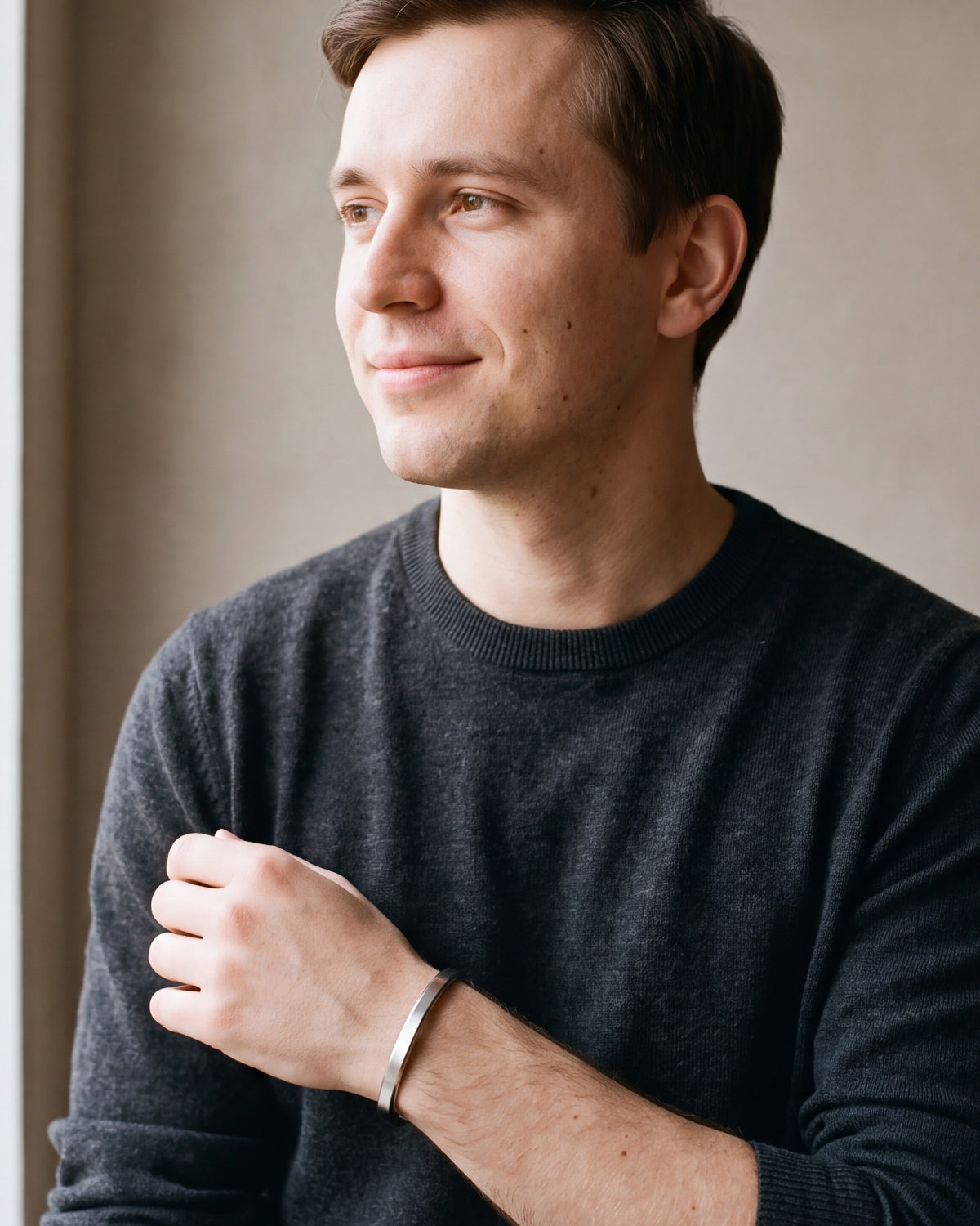 Man in a charcoal sweater looking toward the window, wearing the silver Atlas flat cuff bracelet