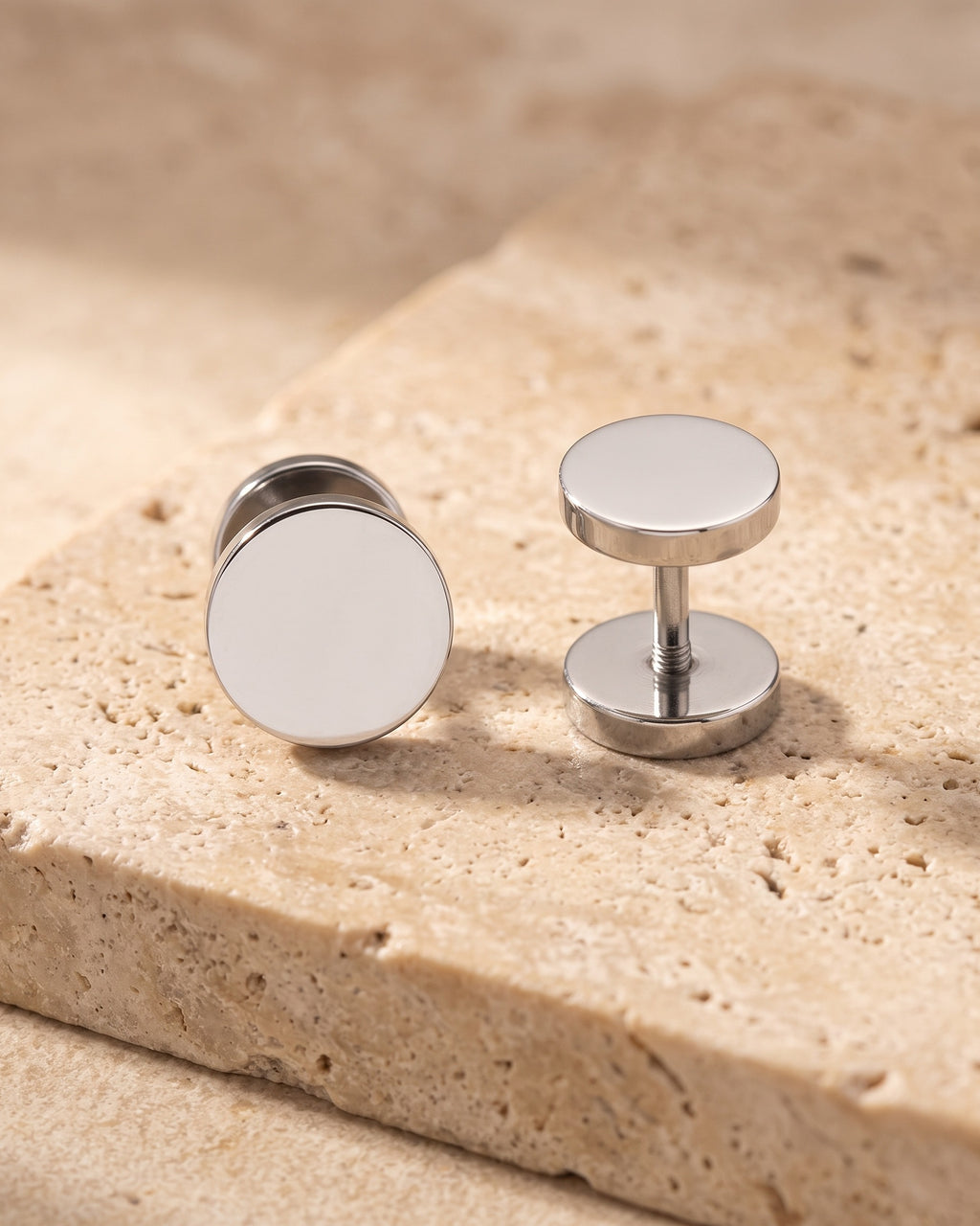 Entire Atlas flat disc barbell stud earrings in silver-tone steel, lying flat on warm stone