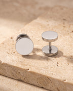 Entire Atlas flat disc barbell stud earrings in silver-tone steel, lying flat on warm stone