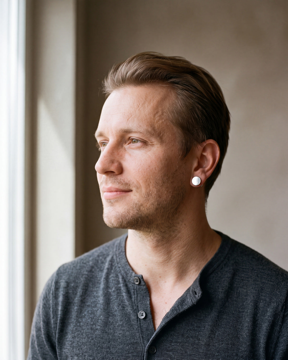 Man in a charcoal knit henley by a window wearing the Atlas flat disc barbell stud earrings in silver-tone steel