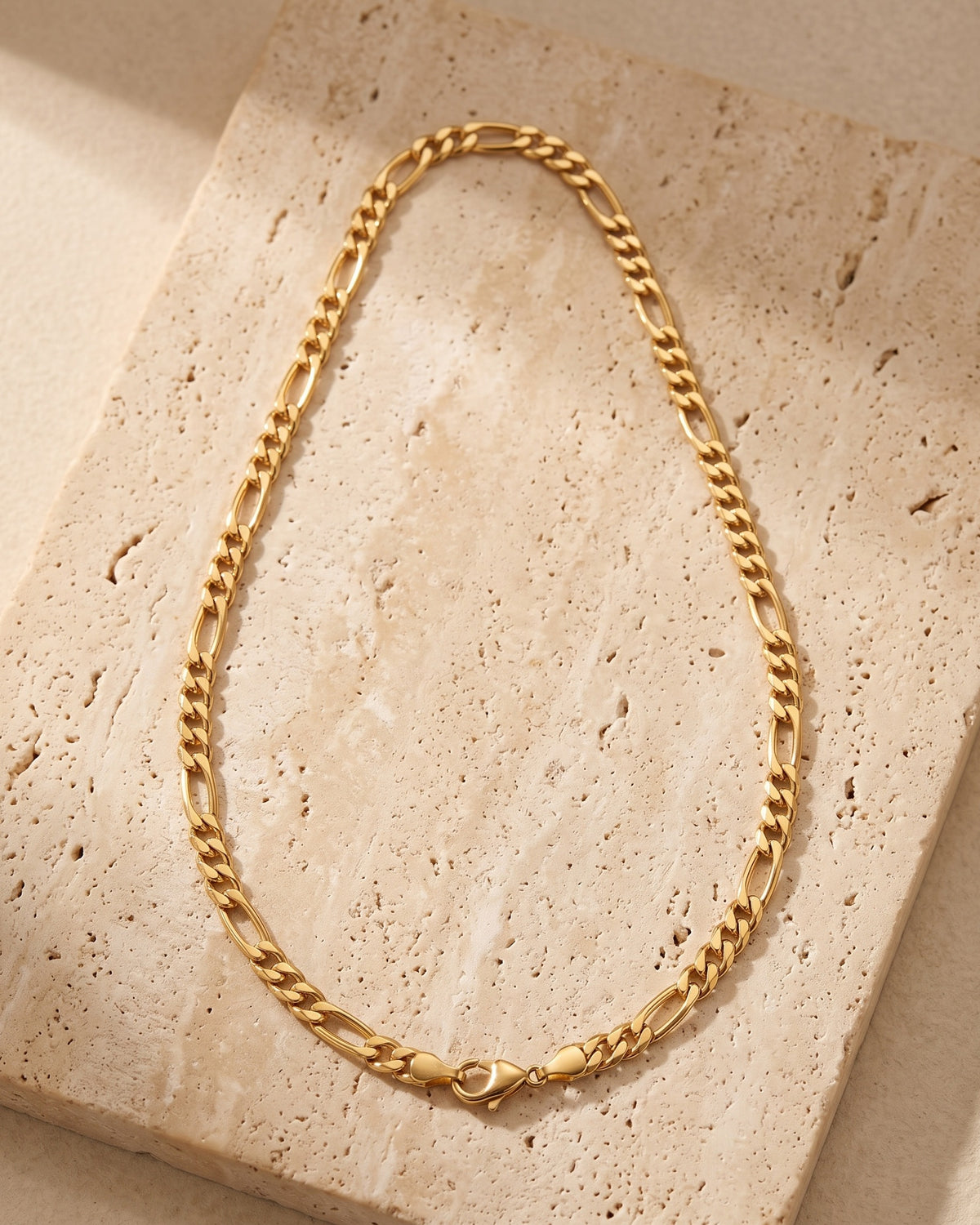 The whole gold-tone Atlas Flat Gold-Tone Figaro Chain lying on warm travertine stone