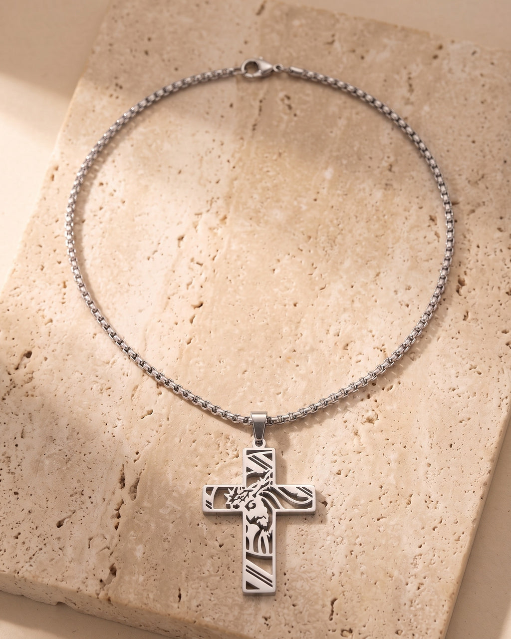 Silver-tone Atlas cut-out cross pendant and box chain laid whole on warm stone